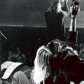 Black and white image of the members of 'Band of Skulls' on stage performing taking from behind them and the crowd is in front with a man in the bottom right with his hand raised in the air.